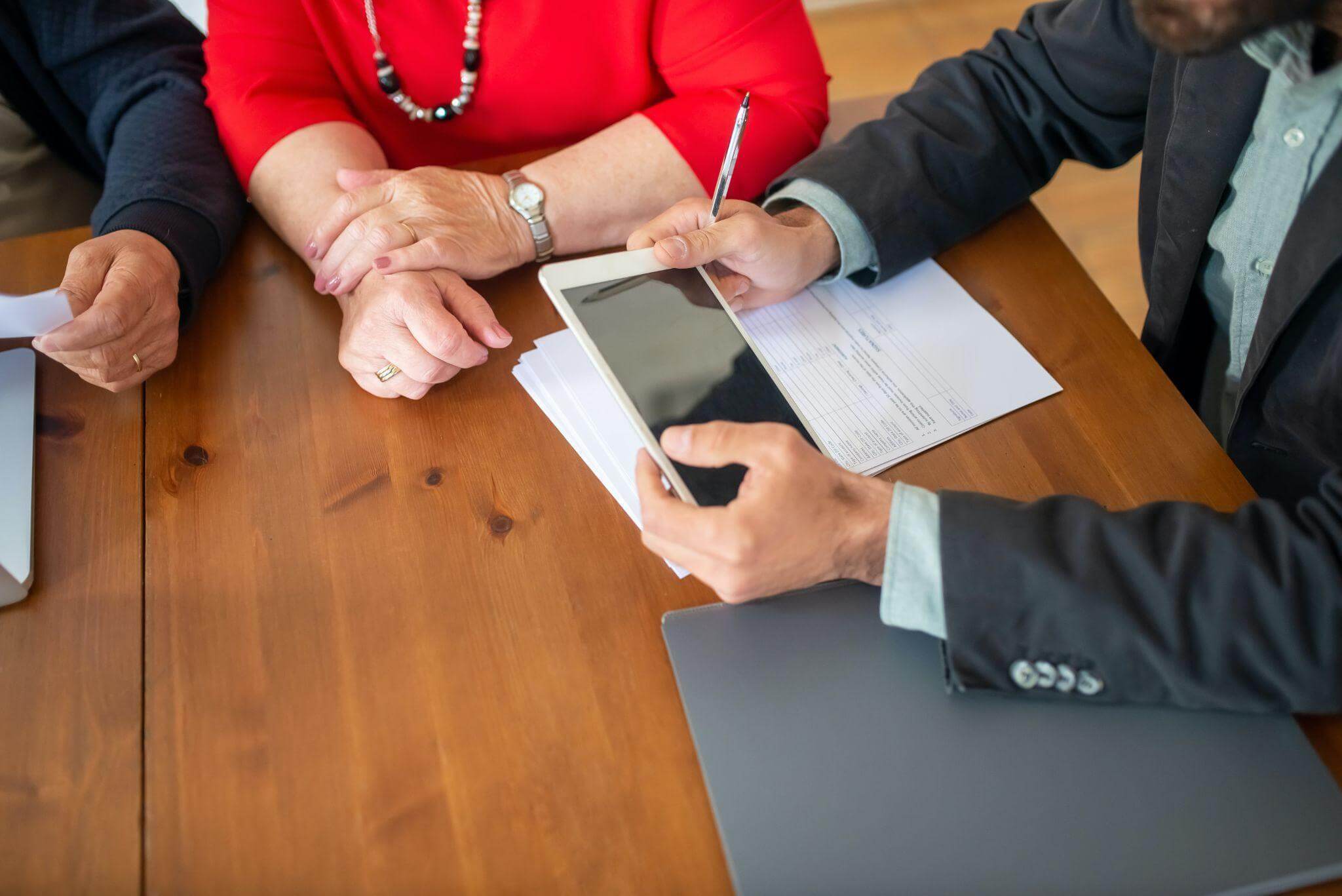 A meeting with three individuals at a table. One person holds a device above papers, while a woman in a red blouse sits next to a person with a laptop