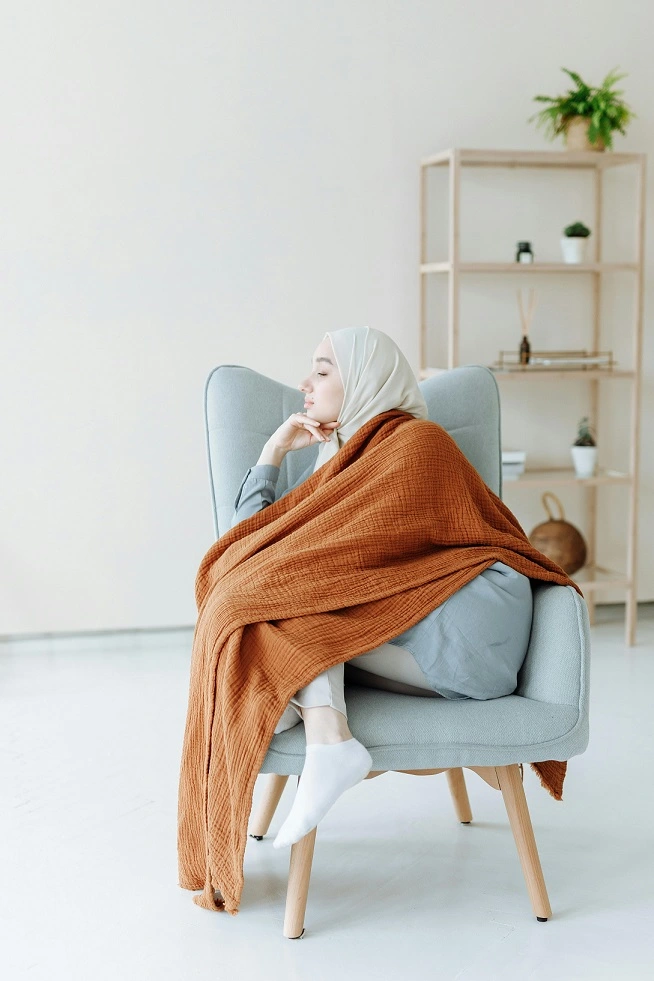 A woman in white hijab sitting on a blue couch covered in an orange blanket.