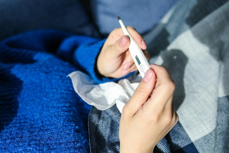 Memeriksa suhu dengan termometer. Seseorang dalam sweater biru dan selimut terbaring dengan tisu di perut.